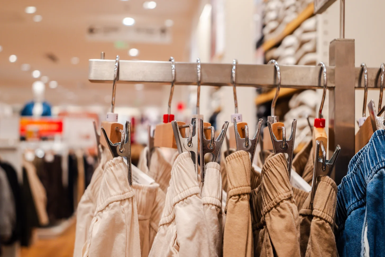 Knitted sweaters in cream, tan, and blue hang from clips on a wooden rod in a retail store, with a blurred shopping area visible in the background.