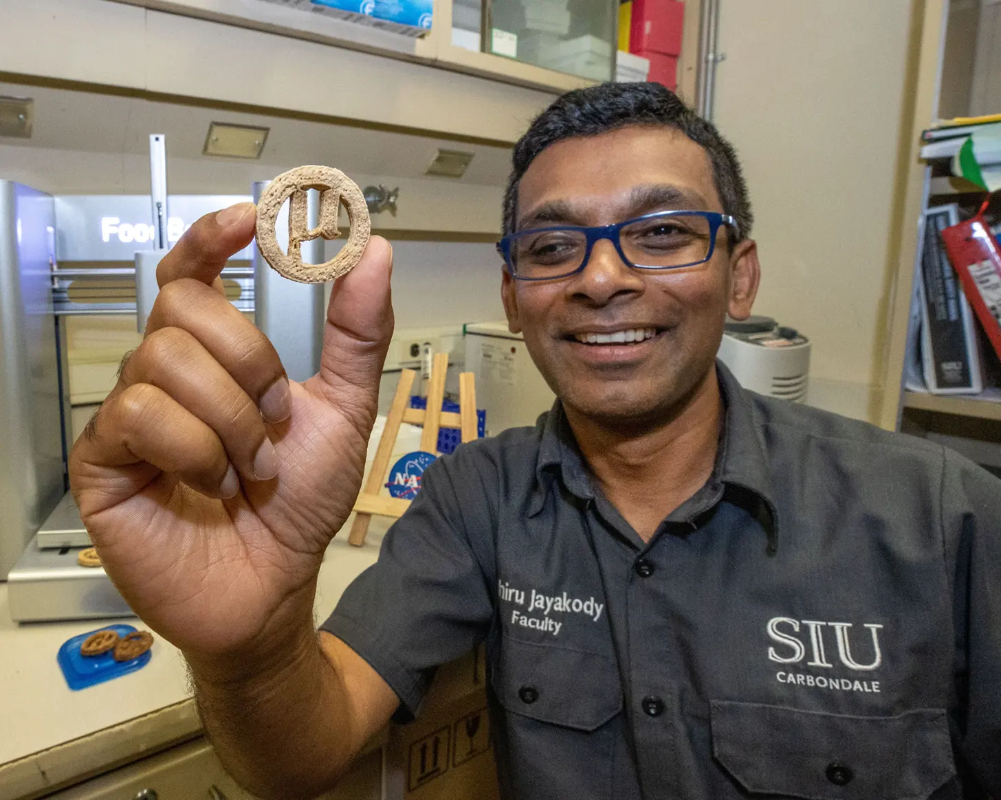 A man wearing glasses and a dark SITU Carbondale Faculty shirt holds up a circular food object with a cutout design in a laboratory setting.