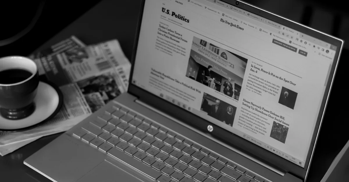 Laptop displaying a New York Times article titled "U.S. Politics" with a cup of coffee and folded newspaper beside it on a dark surface, photographed in black and white.