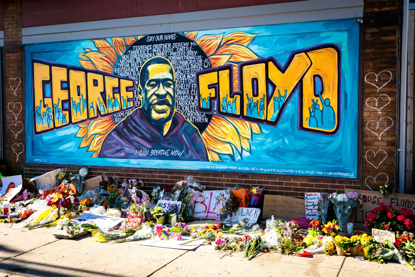Mural on brick wall depicts a portrait of a man with text reading "GEORGE FLOYD" and "I CAN BREATHE NOW" surrounded by golden wings and blue figures. Below the mural, numerous flowers, candles, and handwritten signs cover the sidewalk.