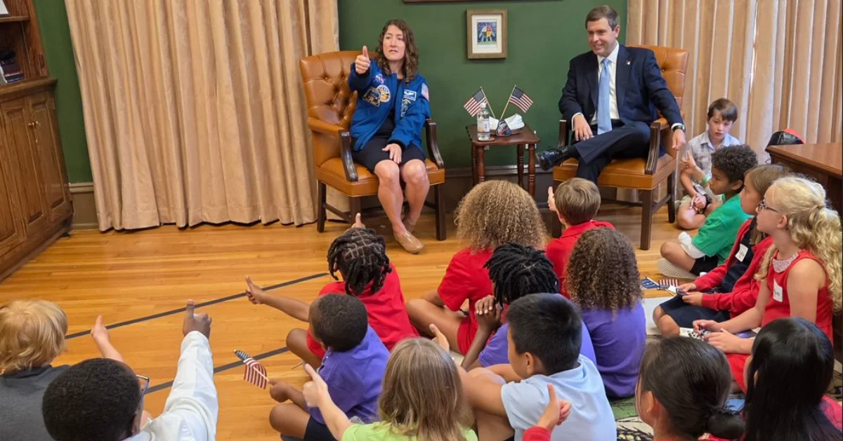 A woman in a blue flight suit sits in a leather chair speaking to a group of children seated on the floor in a wood-paneled room with green walls. A man in a dark suit sits nearby. American flags are visible on a table between them.