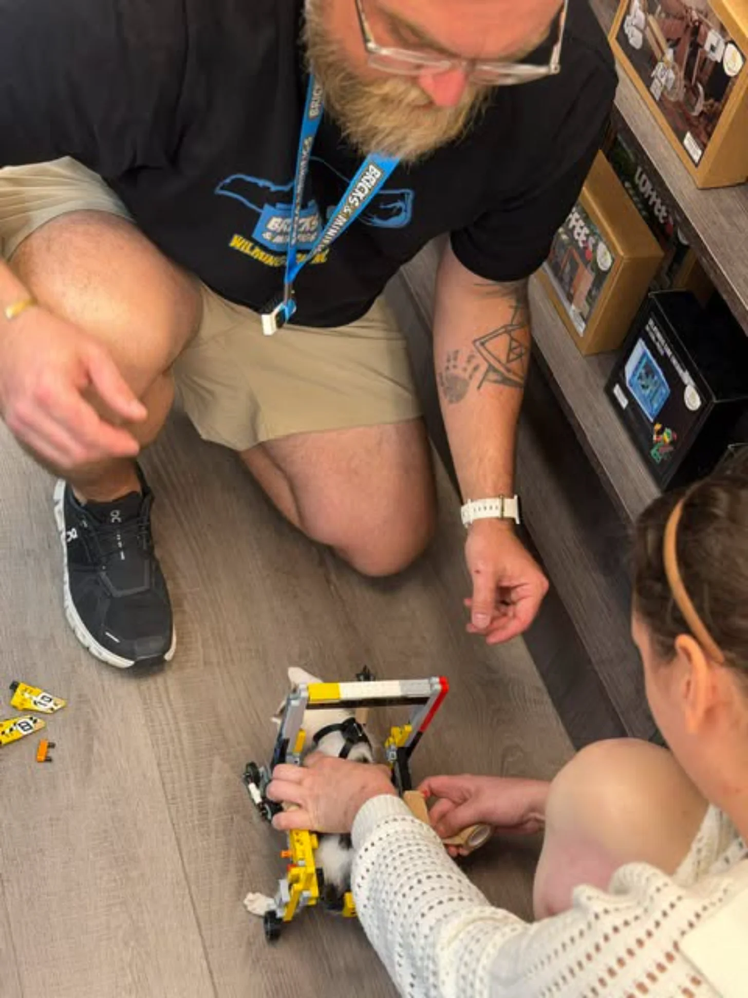 A man wearing glasses and a lanyard kneels on the floor while a woman holds a yellow and black handheld tool over a small kitten. Cardboard boxes are visible in the background.