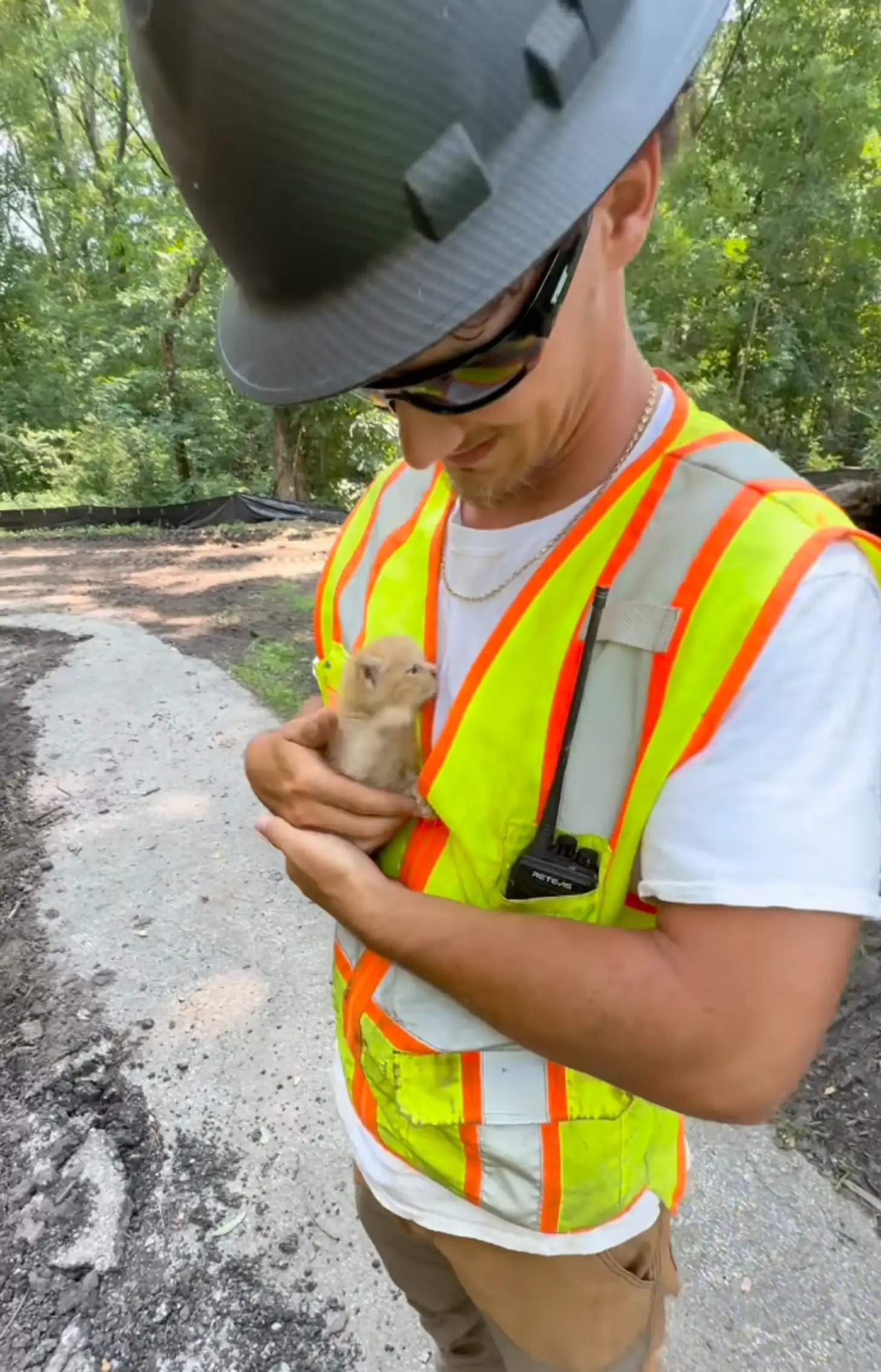 A worker in a gray hard hat, sunglasses, and high-visibility yellow and orange safety vest holds a small tan-colored animal in his hands while standing on a dirt road lined with trees.