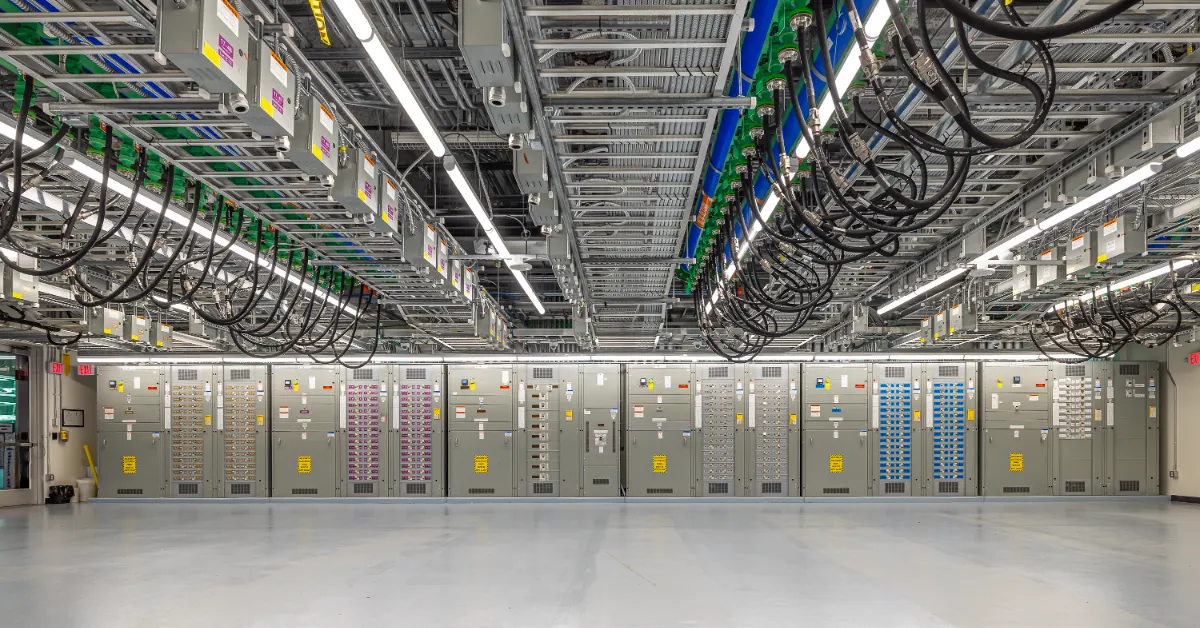 Large electrical distribution cabinets with colored wiring and circuit breakers line a concrete floor beneath an industrial ceiling with cable trays and fluorescent lighting.