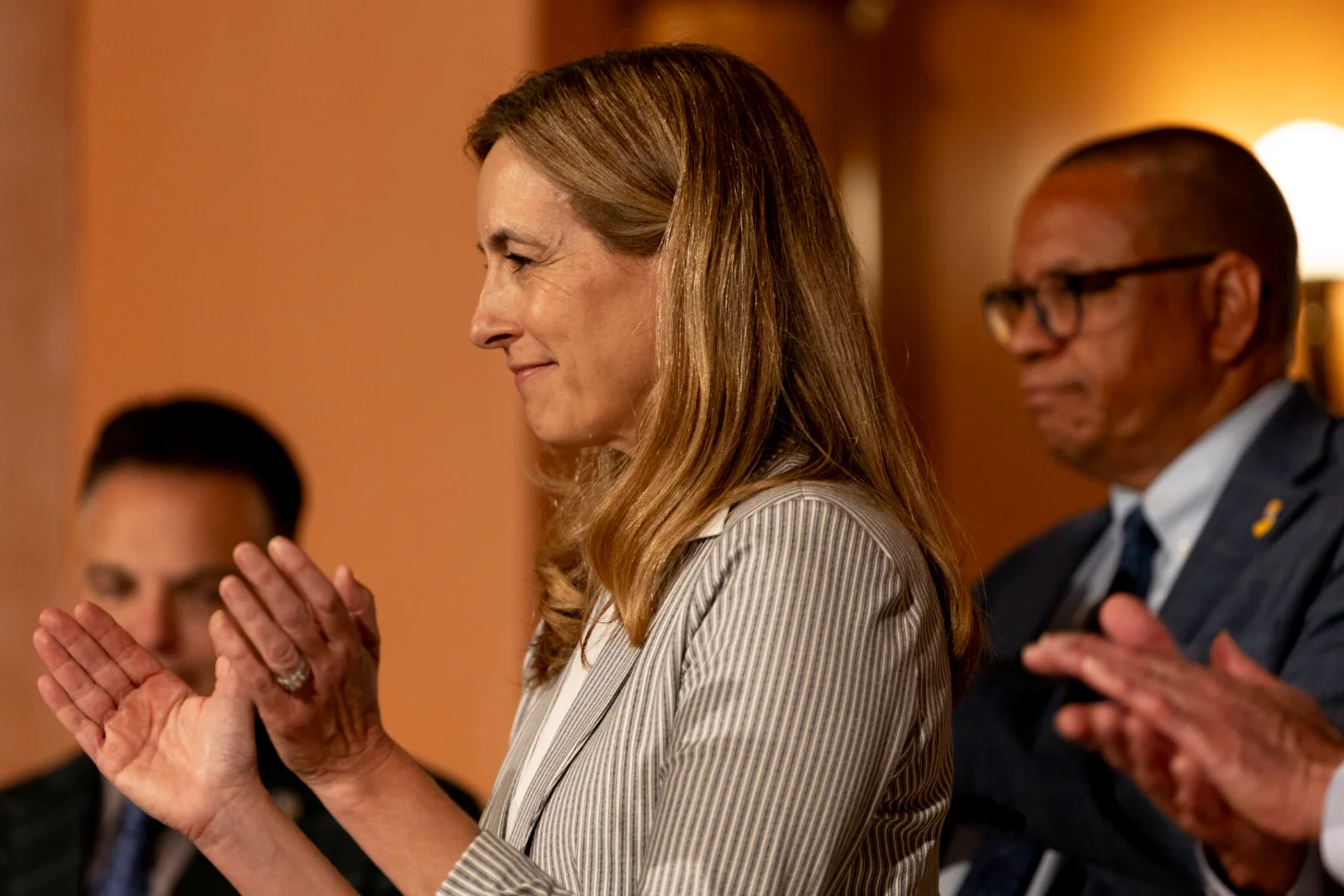 A woman in a gray striped blazer speaks while gesturing with her hands, flanked by two men in dark suits against an orange-toned background.