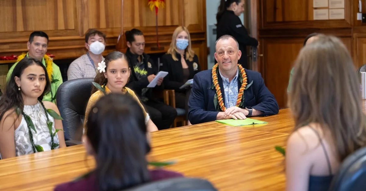 A man in a dark blazer and lei sits at a wooden table facing a woman with long brown hair, in what appears to be a formal indoor setting. Several other people wearing lei stand and sit nearby.