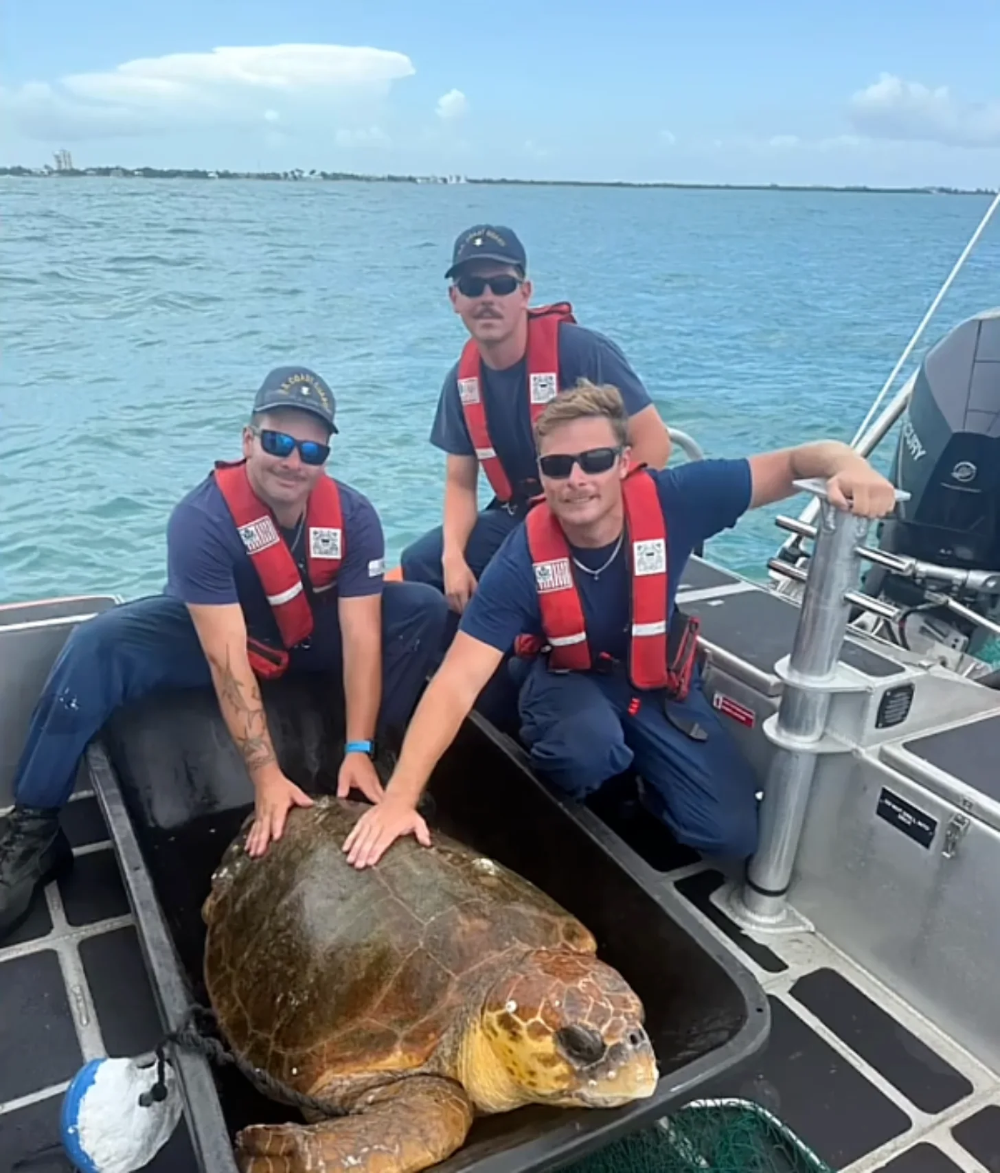 Three men in navy shirts and red life vests pose with a large sea turtle in a boat on open water.