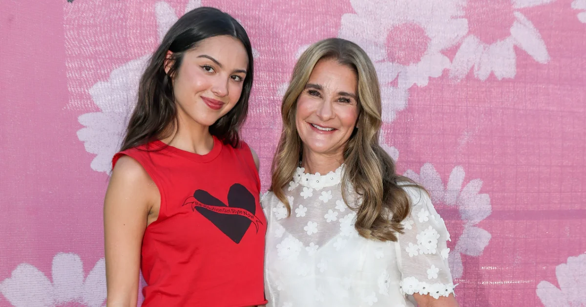 Two women, Olivia Rodrigo and Melinda French Gates, stand together against a pink background with white flower patterns. The woman on the left wears a red sleeveless shirt with a black heart design. The woman on the right wears a white lace top with short sleeves.