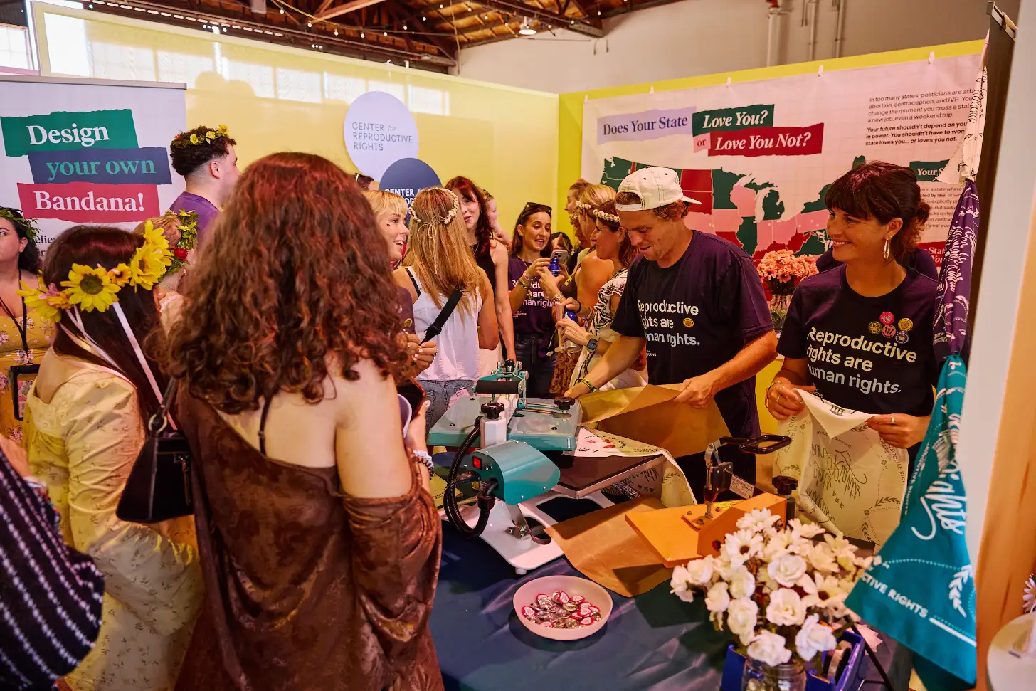 A group of people gather at a booth with a sign reading "Does Your State Love You? or Love You Not?" Two people wearing purple shirts that read "Reproductive rights are human rights" stand behind a table with a bandana-decorating station, including a turquoise machine and flowers in a vase. Visitors interact with the booth while colorful signage and banners are visible in the background.