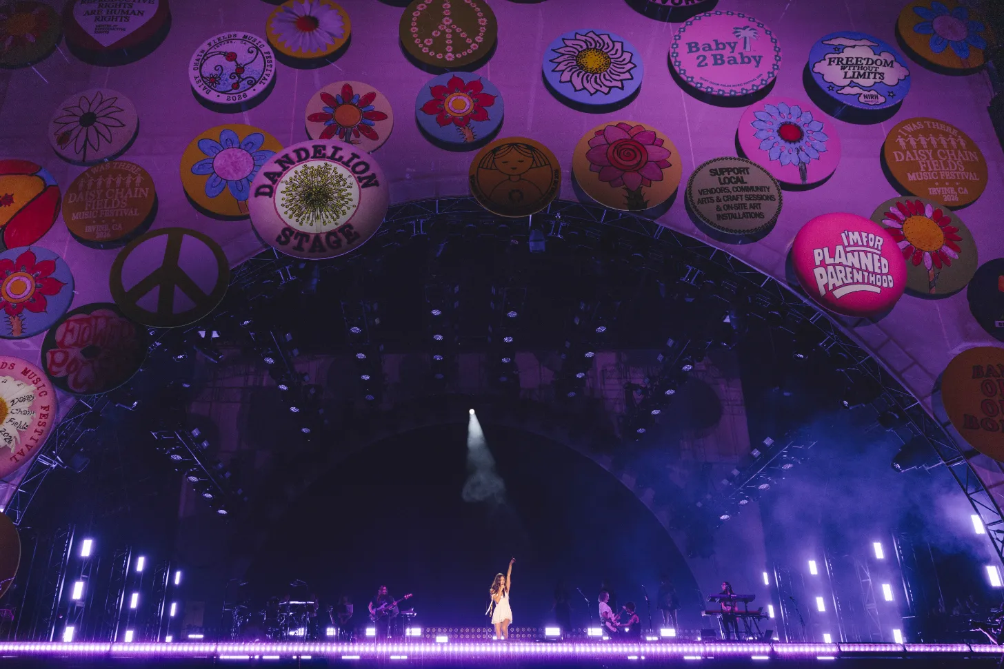 A performer in a white dress stands center stage under bright stage lighting, with band members visible on either side. Large colorful decorated circular badges hang from the purple ceiling above, including designs with flowers, peace symbols, and text reading "Baby2Baby," "Planned Parenthood," and "Dandelion Stage."