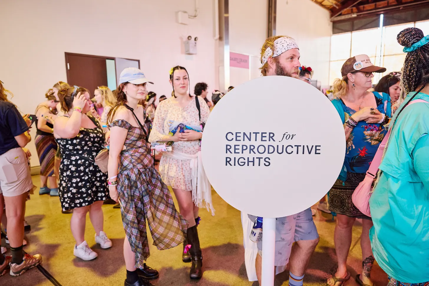 A large crowd of people in colorful outfits stands indoors on a yellow floor. A man in the center holds a large white circular sign that reads "CENTER for REPRODUCTIVE RIGHTS" in dark blue text.