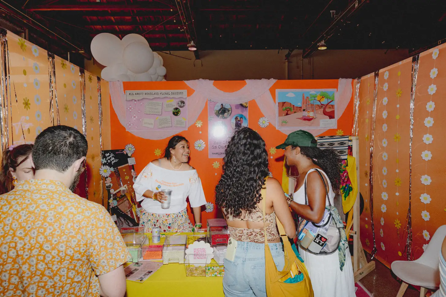 A woman in a white shirt stands at a yellow table inside a booth decorated with orange panels and white flower patterns, speaking to several visitors. A sign reading "ALL ABOUT WOODLAND FLORAL DESIGNS" is visible on the back wall above displayed images and materials.