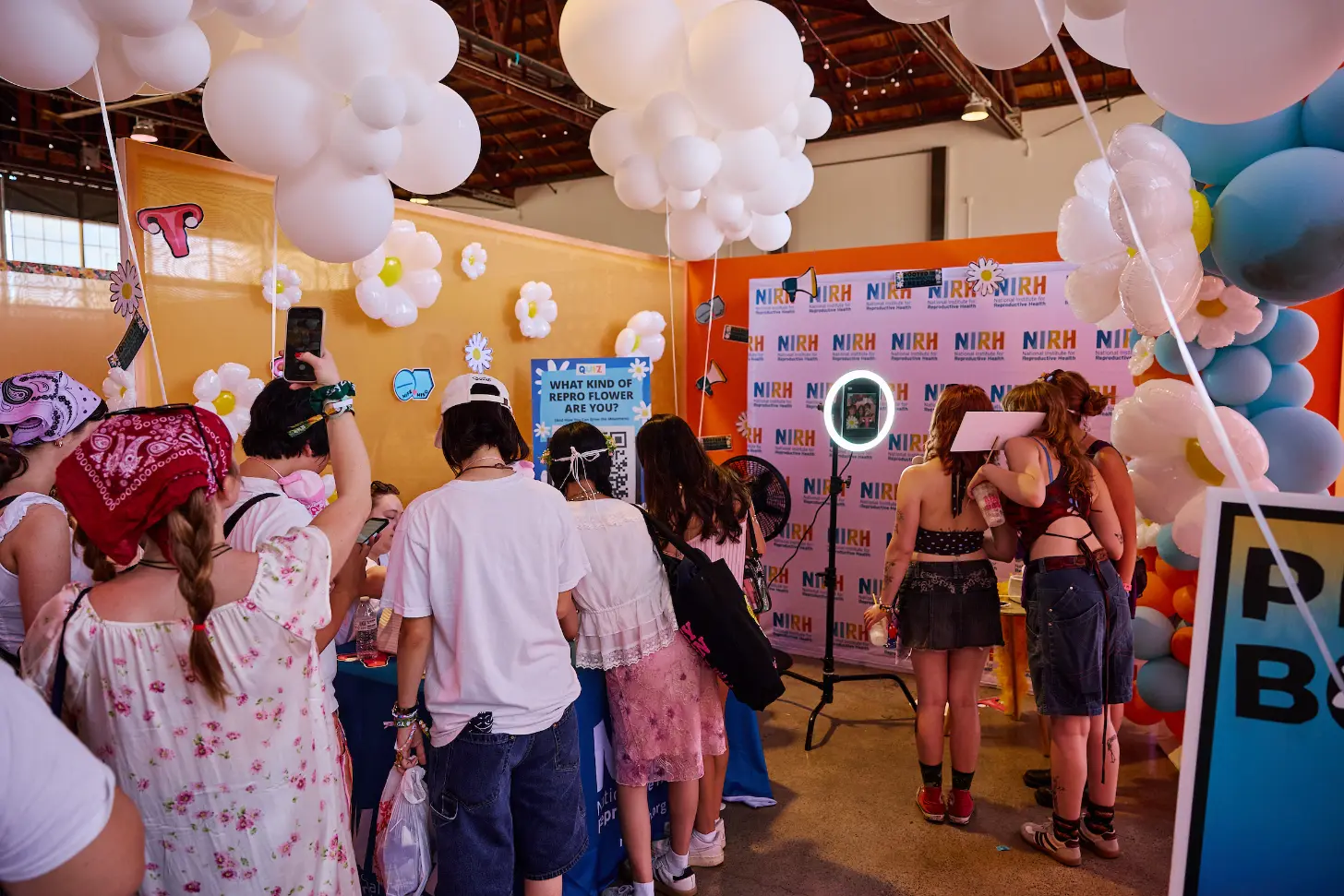A group of people gather in a decorated indoor space with pink and white balloons hanging from the ceiling and white daisy decorations on an orange wall. A backdrop with "NIRH" repeated across it stands behind attendees, some of whom hold phones to take photos. A sign reads "WHAT KIND OF REPRO FLOWER ARE YOU?"