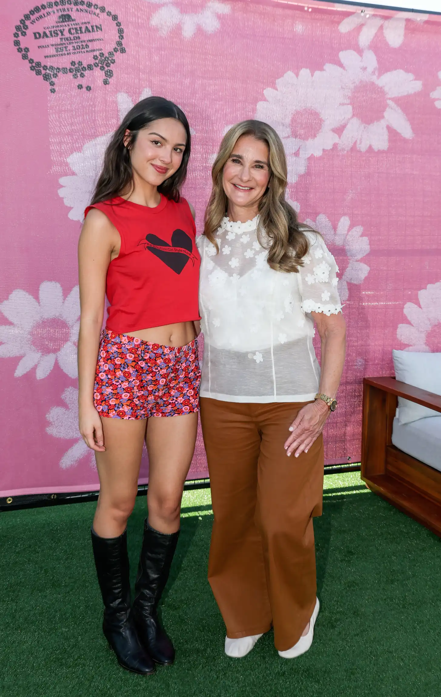 Two women, Olivia Rodrigo and Melinda French Gates, stand together against a pink background with white flower patterns. The woman on the left wears a red sleeveless shirt with a black heart design. The woman on the right wears a white lace top with short sleeves.