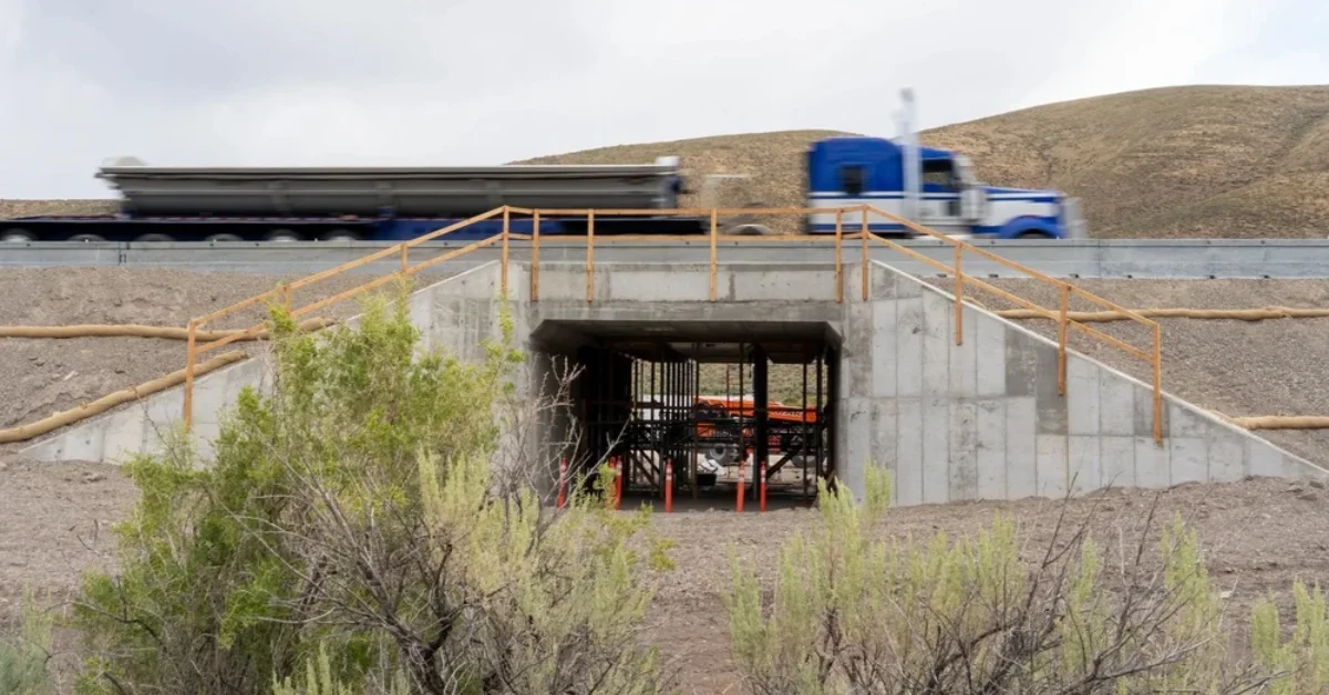 A concrete underpass with yellow metal railings sits in an arid landscape. A blue and white semi truck with an empty flatbed trailer is visible on the road above the structure. Sparse green shrubs grow in the foreground.