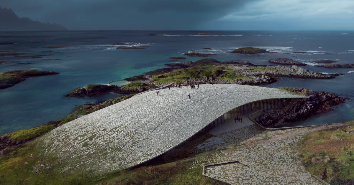 A large curved stone structure sits on a rocky island surrounded by turquoise water and smaller vegetated islands. People stand on the stone surface and near a wooden fence on the structure's right side.