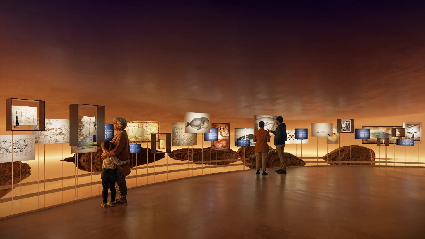 Museum gallery with warm orange lighting displays artwork and photographs on walls and pedestals. Several visitors stand and walk through the space viewing the exhibits.