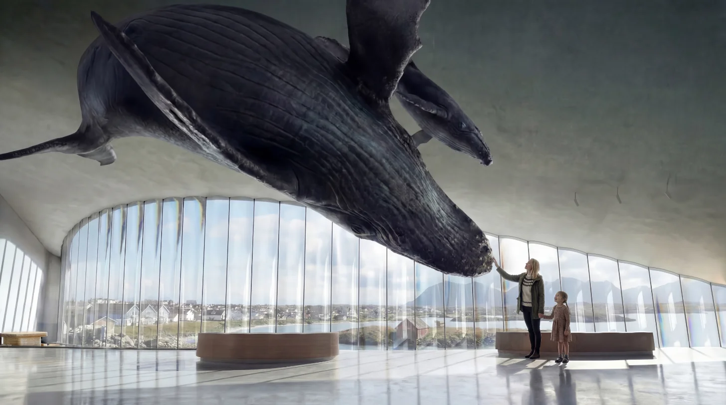 A large dark whale sculpture hangs from the ceiling of a modern building with floor-to-ceiling windows. A woman and child stand below, looking up at the installation. A coastal landscape is visible through the windows.