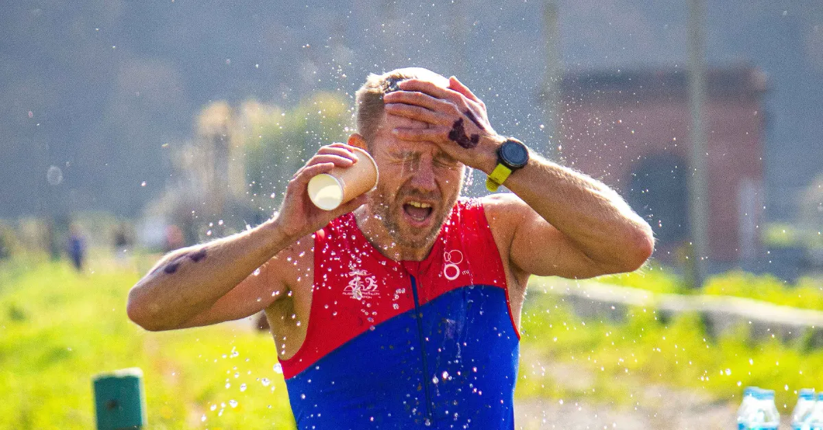 A man wearing a red and blue sleeveless shirt splashes water on his face with both hands, squinting and smiling. Water droplets spray around his head as he holds a paper cup in one hand.
