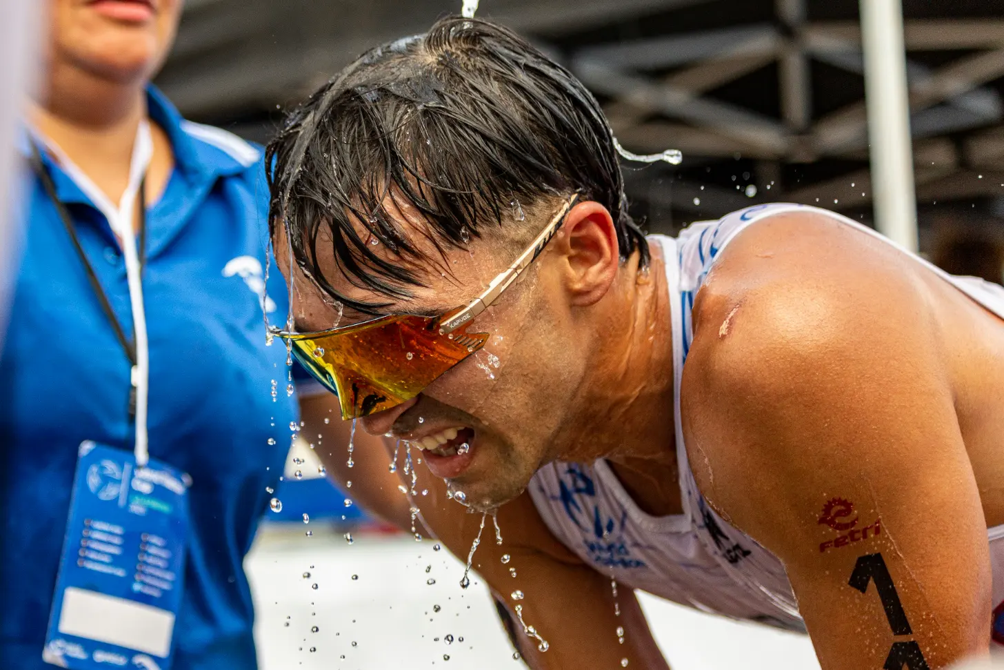 A wet athlete wearing amber-tinted sunglasses and a light-colored tank top wipes water from his face. A person in a blue jacket stands nearby observing.