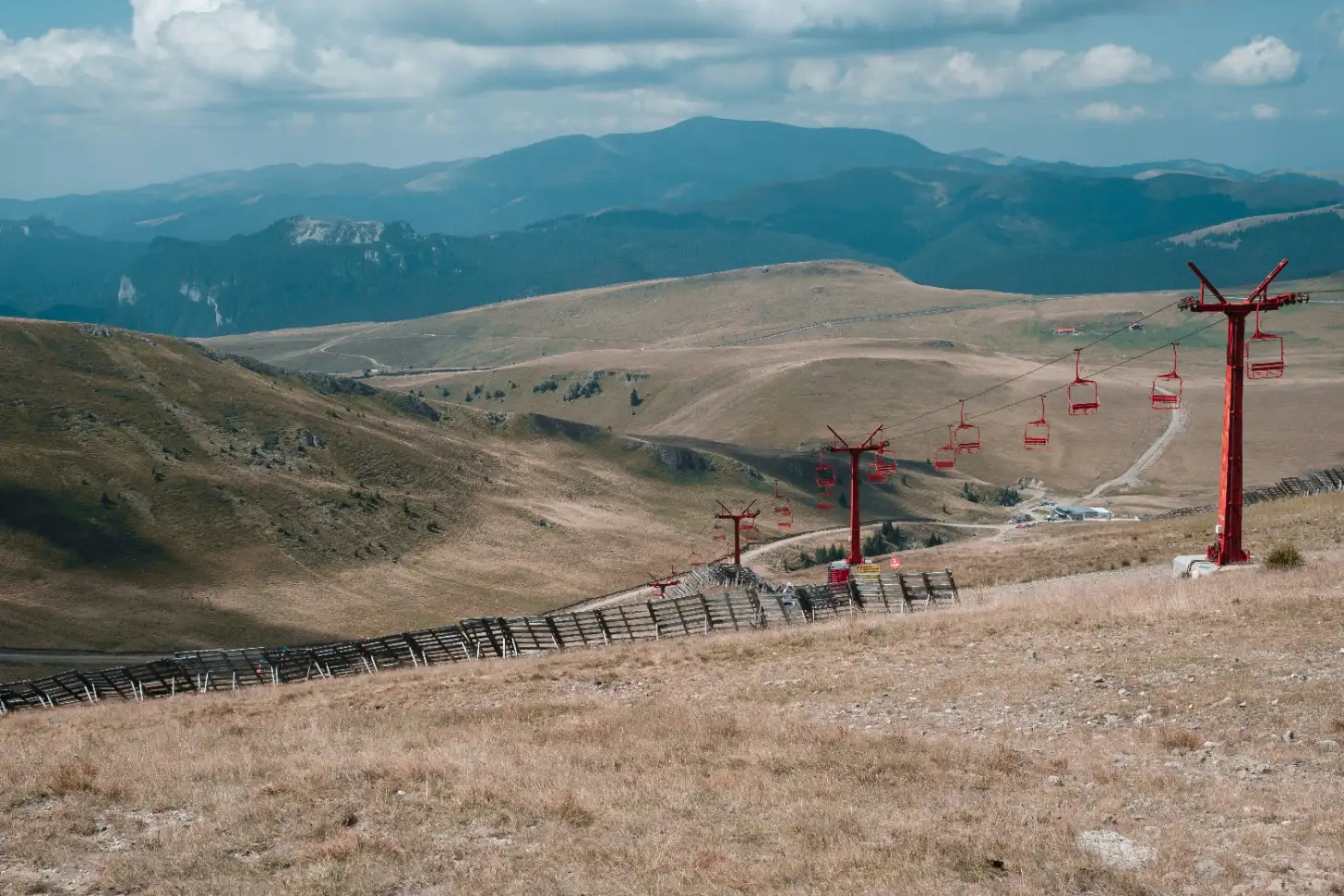 Red ski lift towers and chairs span across a brown, grassy mountainside with forested mountains visible in the distance under a partly cloudy sky.