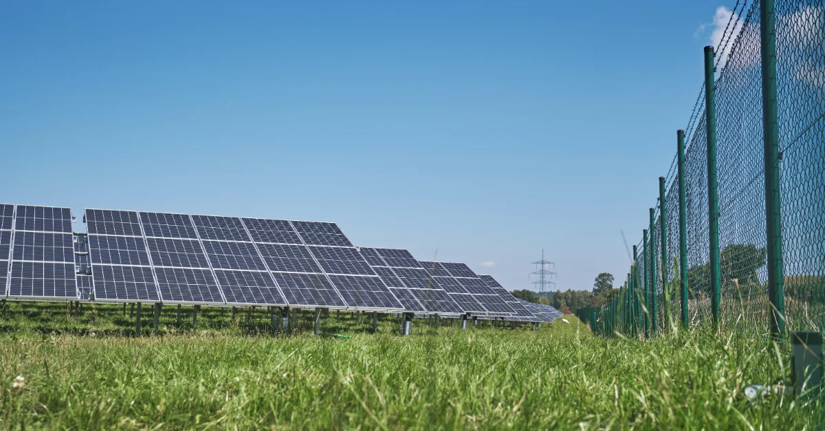 Solar panels mounted on ground-level racks in a grassy field, with a green metal fence visible on the right and electrical transmission towers in the distance under clear sky.