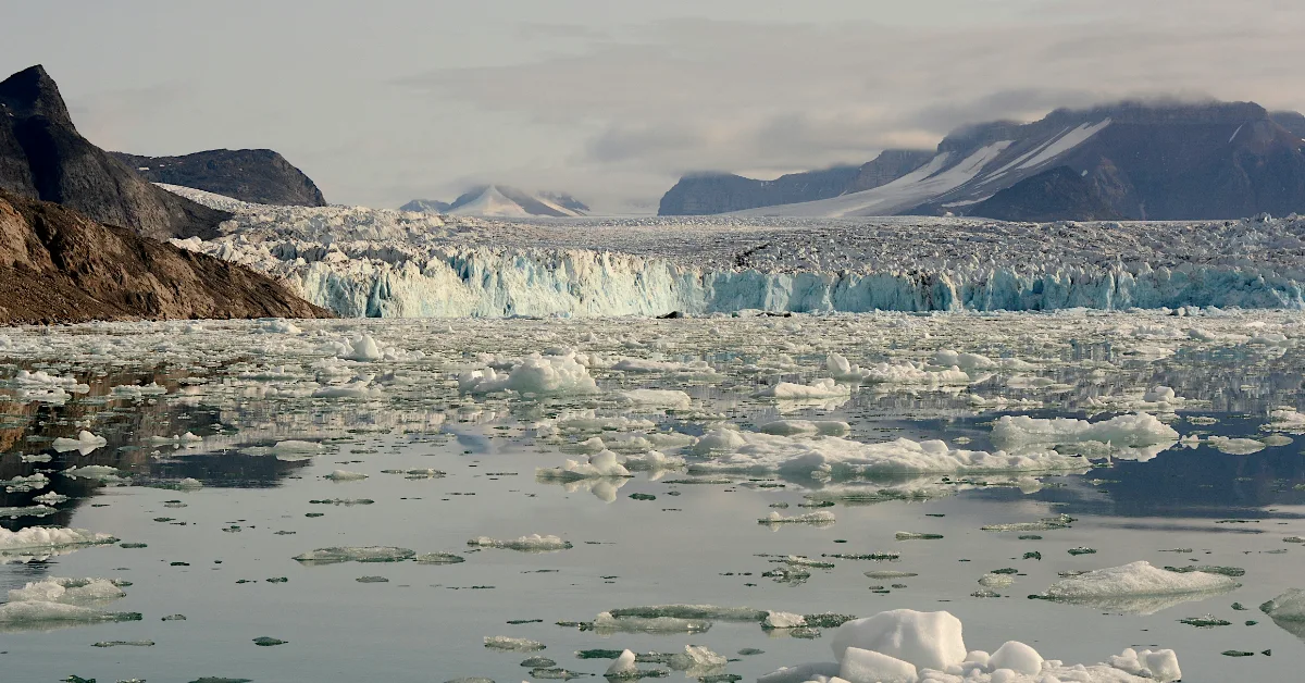Massive glacier with jagged ice cliffs rises behind a bay filled with floating white and blue-green icebergs and ice chunks. Snow-covered mountains visible in the background under a cloudy sky.