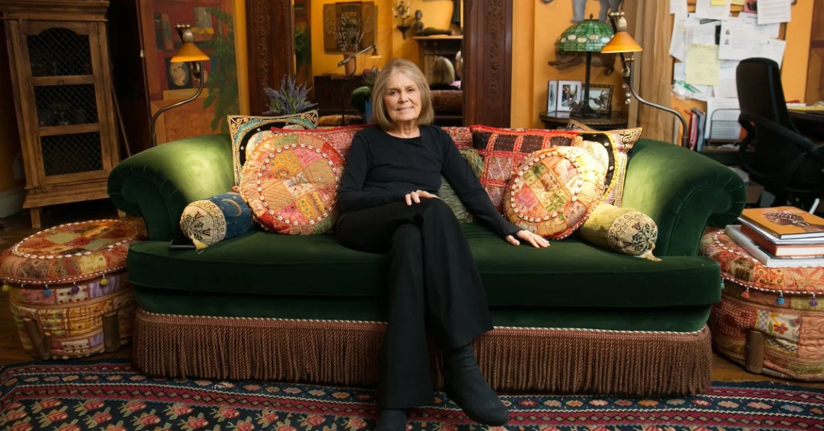 A woman, Gloria Steinem, in black clothing sits on a green upholstered sofa decorated with ornate red and gold patterned pillows in a room with yellow-orange walls, wood furnishings, and decorative objects including lamps and photographs.