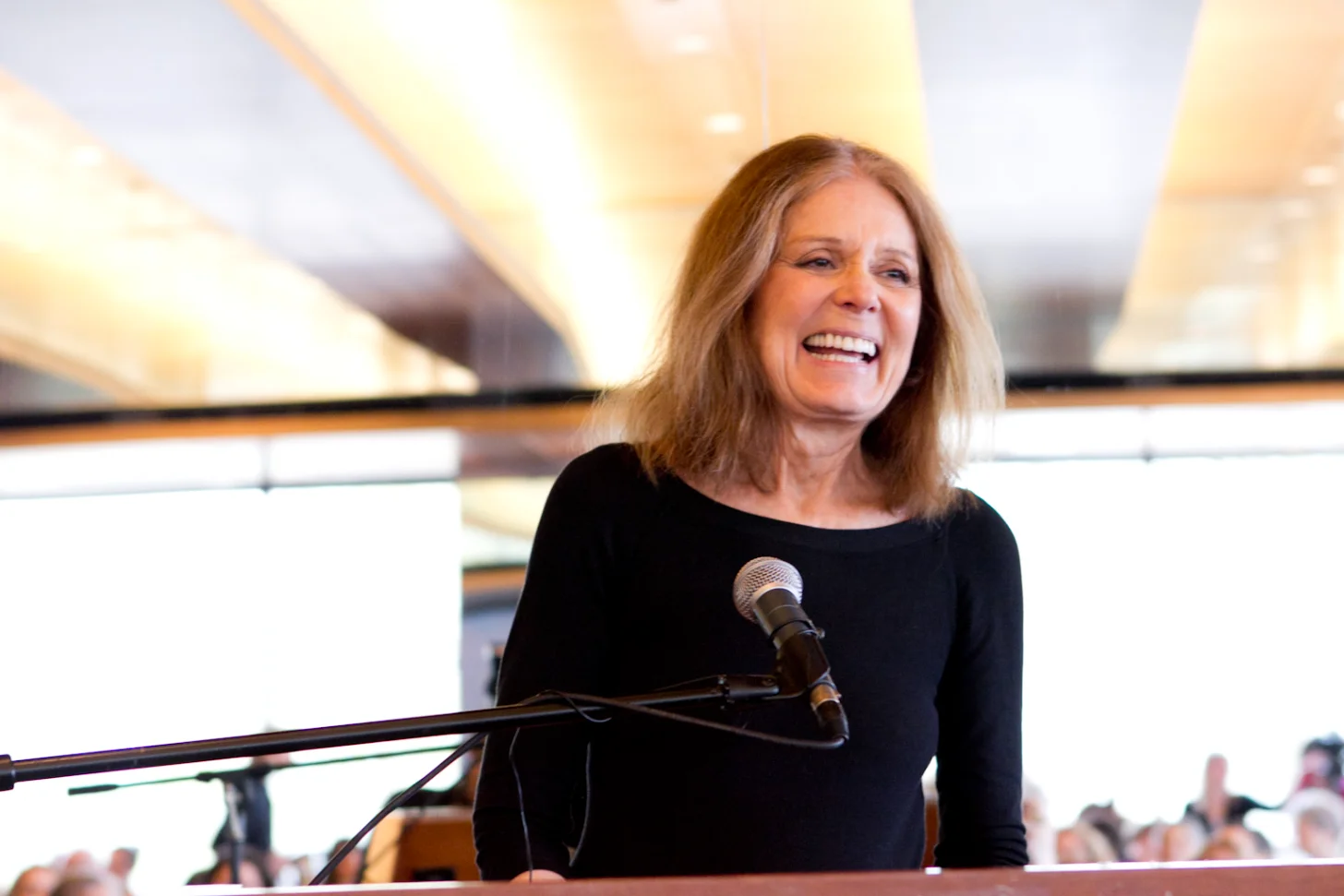 A woman in a black long-sleeved shirt, Gloria Steinem, speaks at a microphone, smiling. She stands before an audience in what appears to be an indoor venue with warm lighting visible in the background.