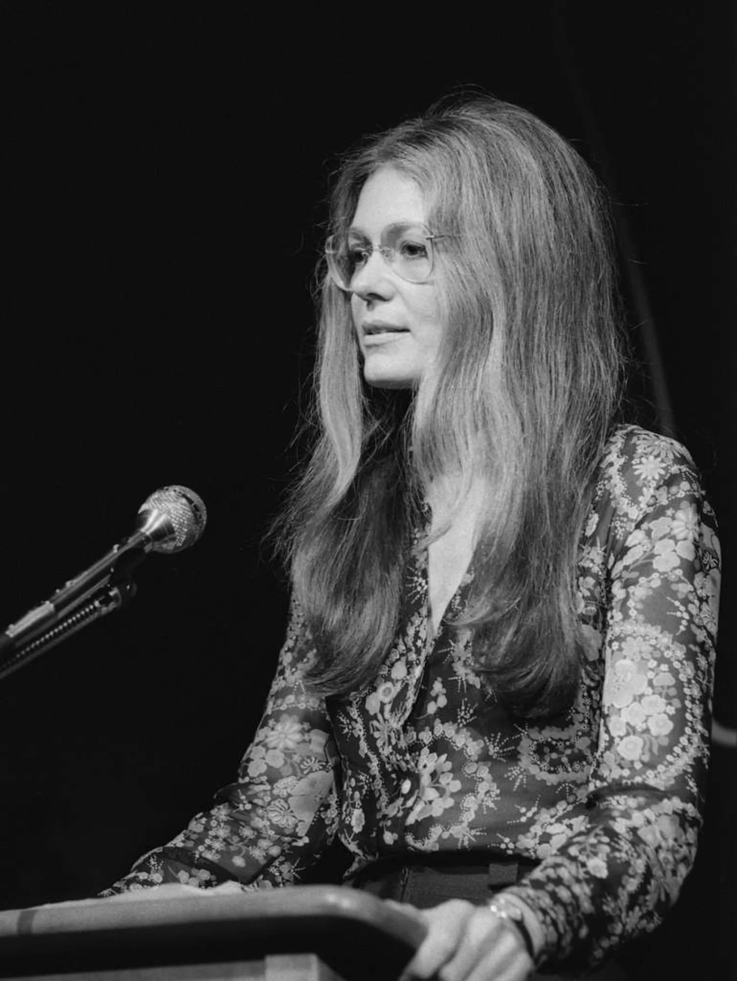 Gloria Steinem, a woman with long hair and glasses speaking at a microphone against a black background, wearing a floral patterned jacket.