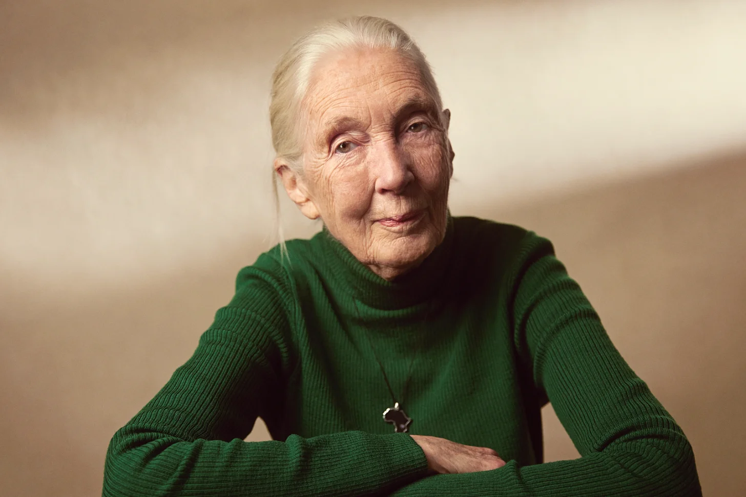Jane Goodall. Elderly woman with white hair wearing a green turtleneck sweater, arms crossed, looking toward the camera against a neutral beige background.