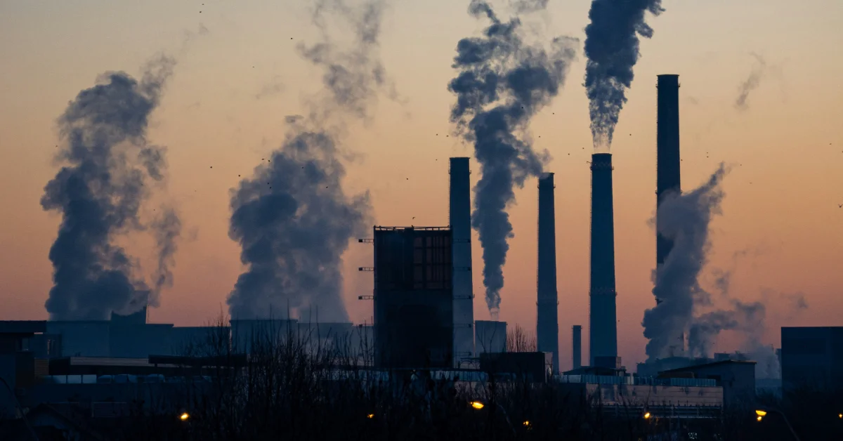 Industrial facility with multiple tall smokestacks emitting dark smoke against a sunset sky at dusk. Silhouetted factory buildings and structures visible in the foreground with lights glowing along the base.
