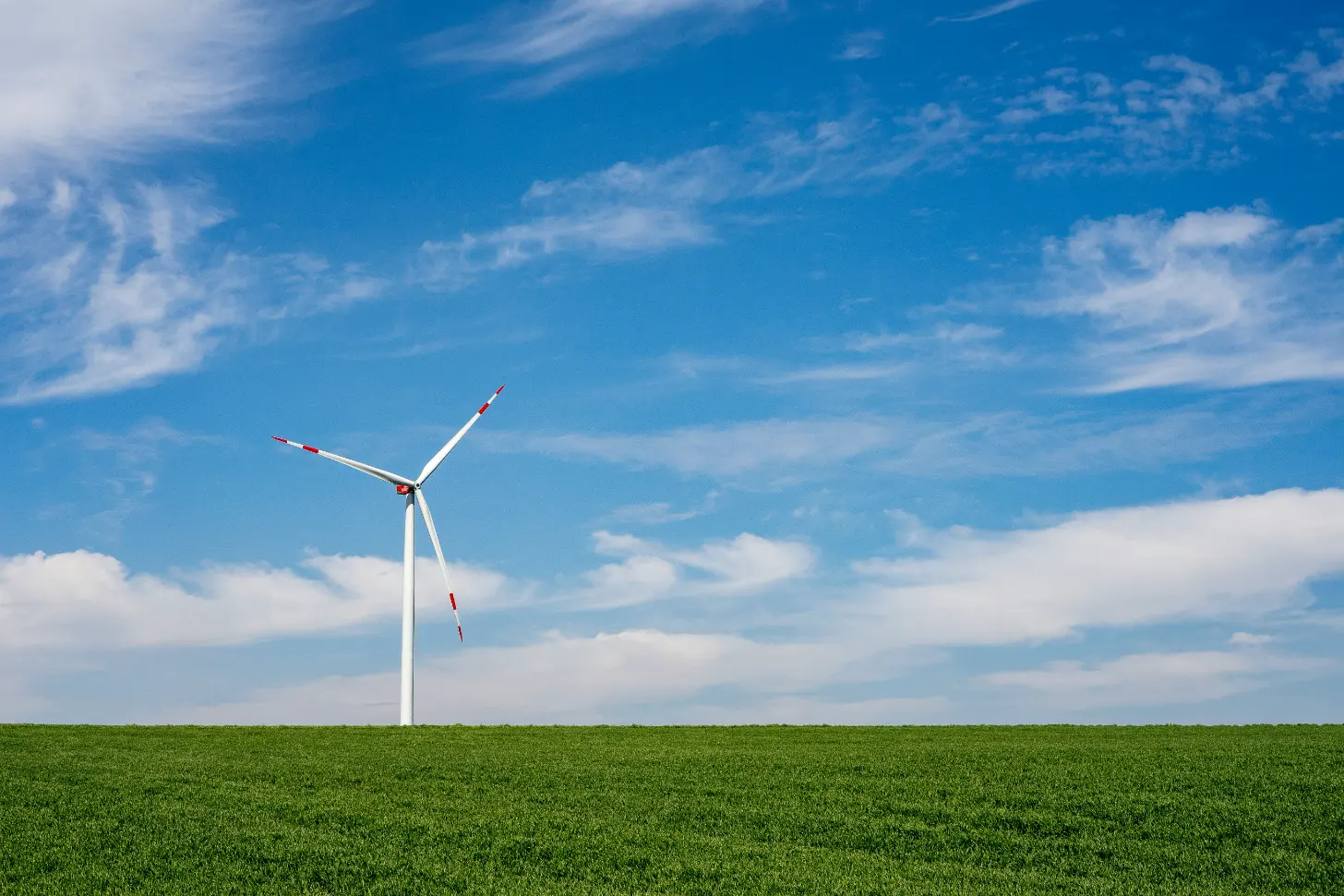 A wind turbine with red and white markings stands in a green field under a blue sky with white clouds.