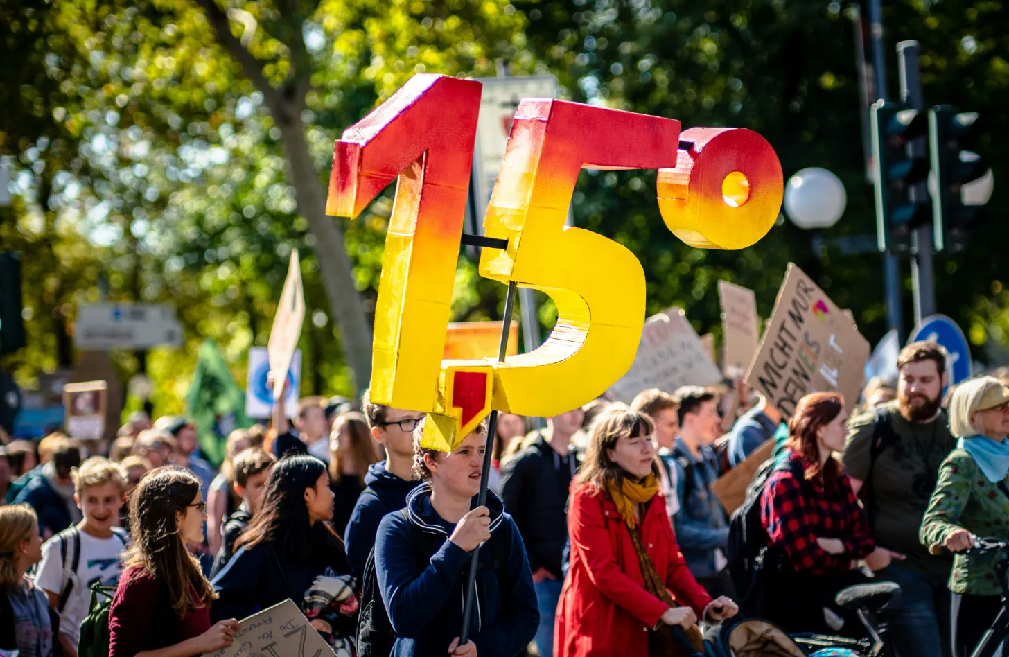 A large yellow and red number "1.5" sign is held by a person in a crowd of protesters gathered outdoors among trees. Several other people holding signs are visible around them.