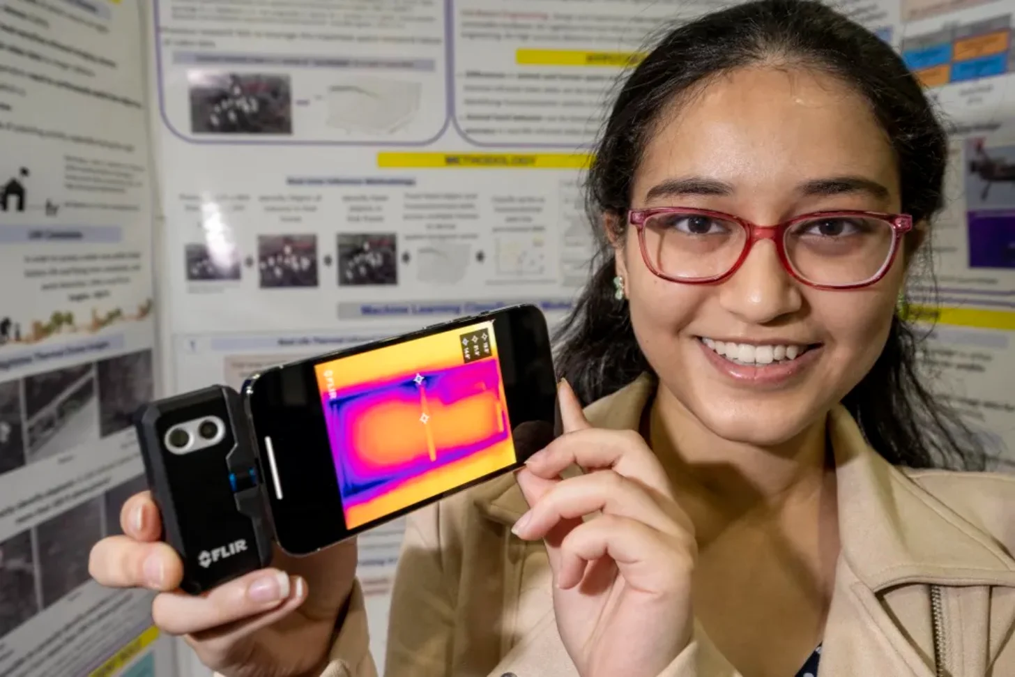 Woman wearing red glasses holds a thermal imaging camera displaying a colorful heat map on its screen, standing in front of research posters.