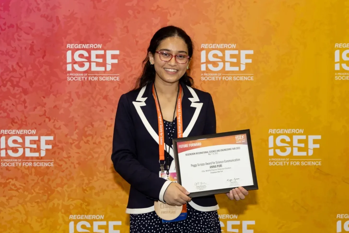 A person wearing glasses and a dark blazer with white trim holds a framed certificate and wears a medal on an orange lanyard in front a Regeneron ISEF backdrop. The certificate reads "Peggy Scripps Award for Science Communication" and "ANIKA PURI."