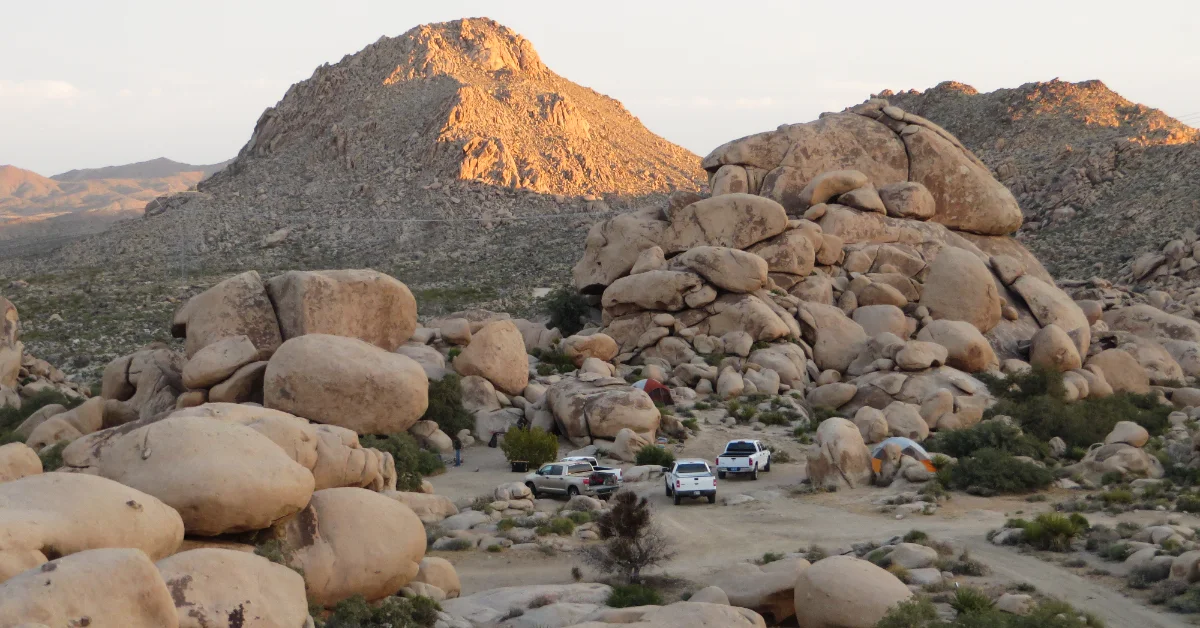 A desert landscape with large rounded boulders in the foreground and a distinctive peaked mountain in the background at sunset. Several vehicles and a tent are parked in a sandy clearing among the rocks.