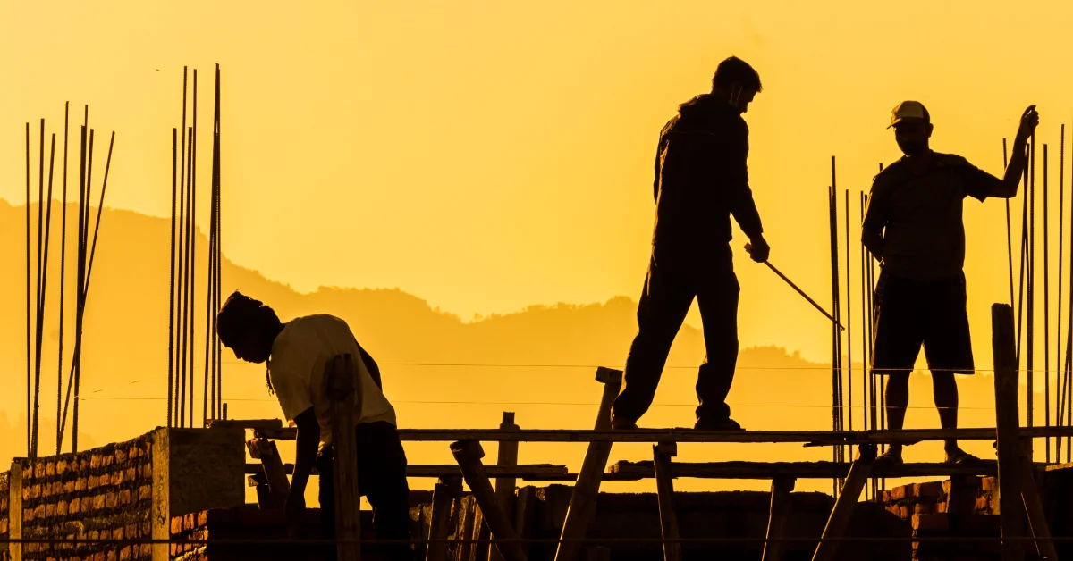 Three construction workers silhouetted against a yellow sky work on a concrete structure with reinforcing steel rods protruding upward; one bends over materials on the left, two others stand on horizontal beams in the center and right of the frame.