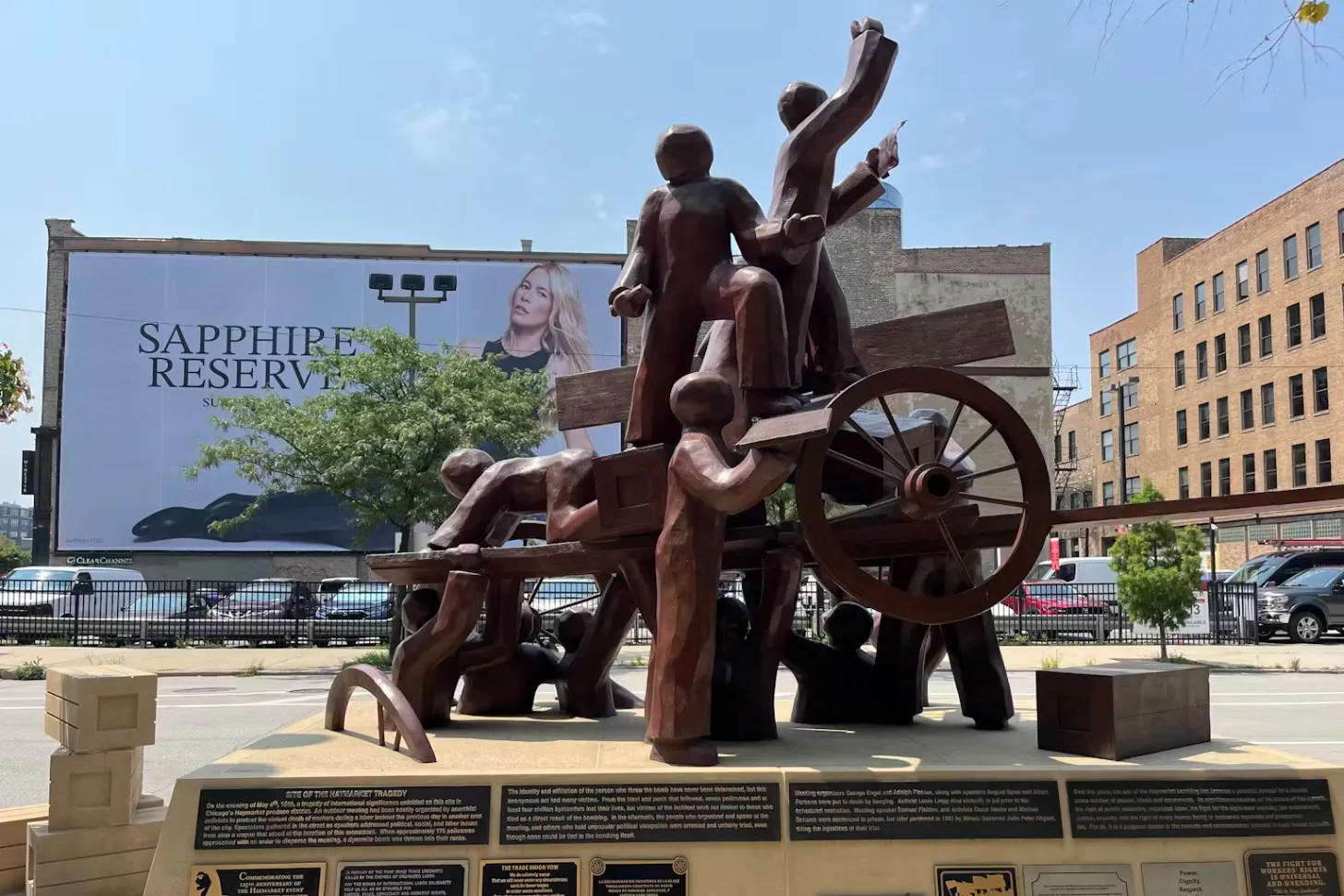 Large bronze sculpture of multiple human figures in dynamic poses on a wooden structure with a wagon wheel, located in an urban plaza with brick buildings and a billboard advertising Sapphire Reserve in the background. Plaques with text are mounted on the base of the monument.