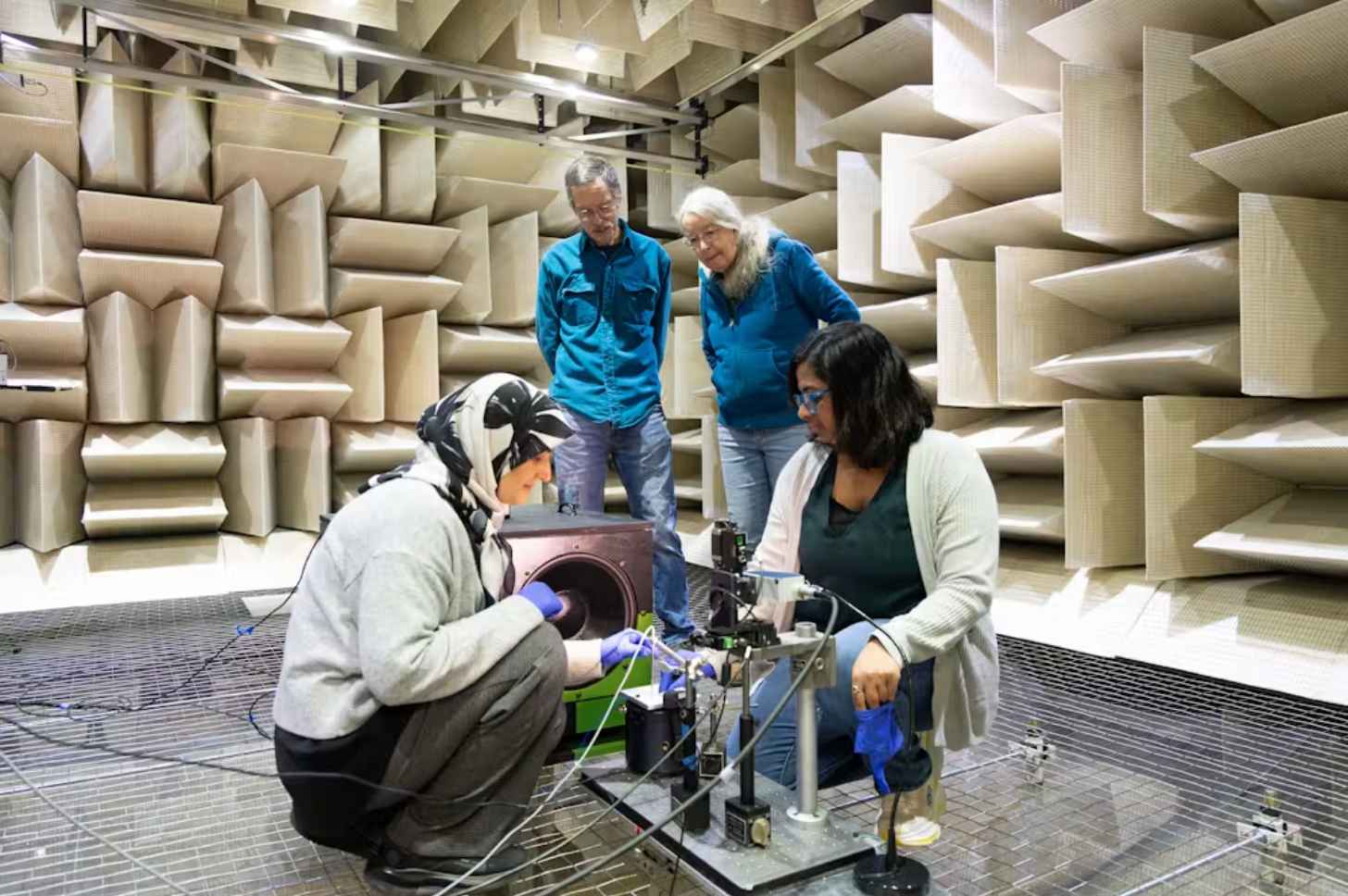 Four people, two standing and observing and two crouching on the ground next to a mechical platform, standing in a chamber with foam tiles covering the walls