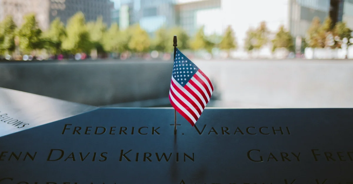 American flag planted on a dark memorial wall engraved with names including "Frederick T. Varacchi" and "Glenn Davis Kirwin," with an urban plaza and trees visible in the background.