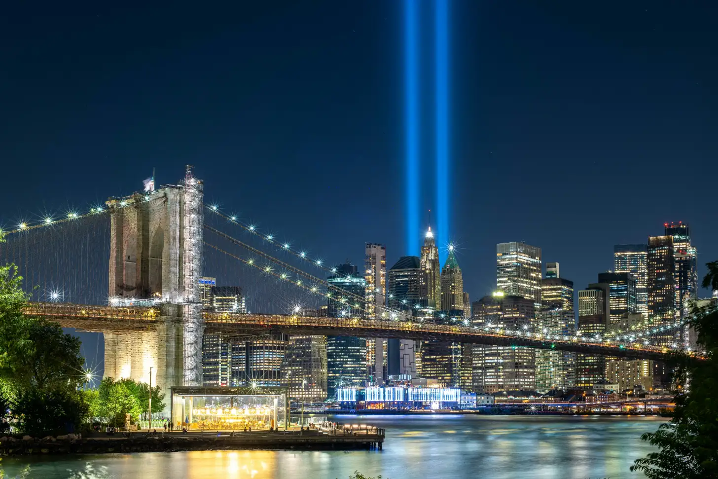 Brooklyn Bridge illuminated at night with Manhattan skyline in background. Two blue light beams shine upward from lower Manhattan as the East River reflects the lit bridge and buildings below.