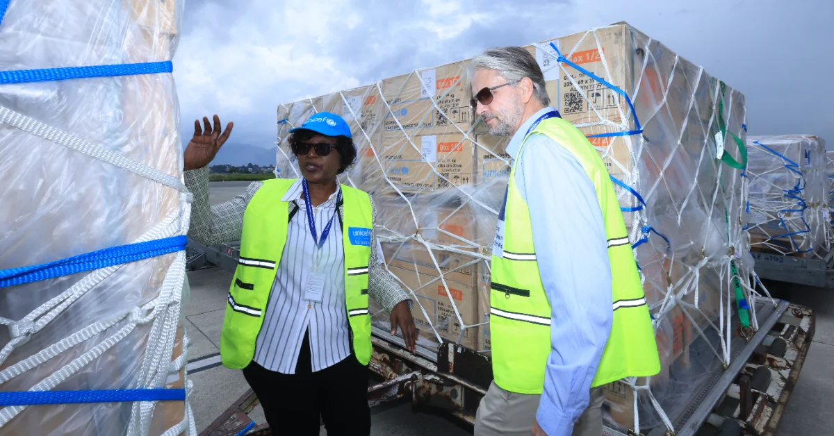 Two workers in yellow safety vests and hard hats stand beside large cardboard boxes wrapped in plastic and blue strapping on what appears to be an airport or cargo tarmac, with an overcast sky visible in the background.
