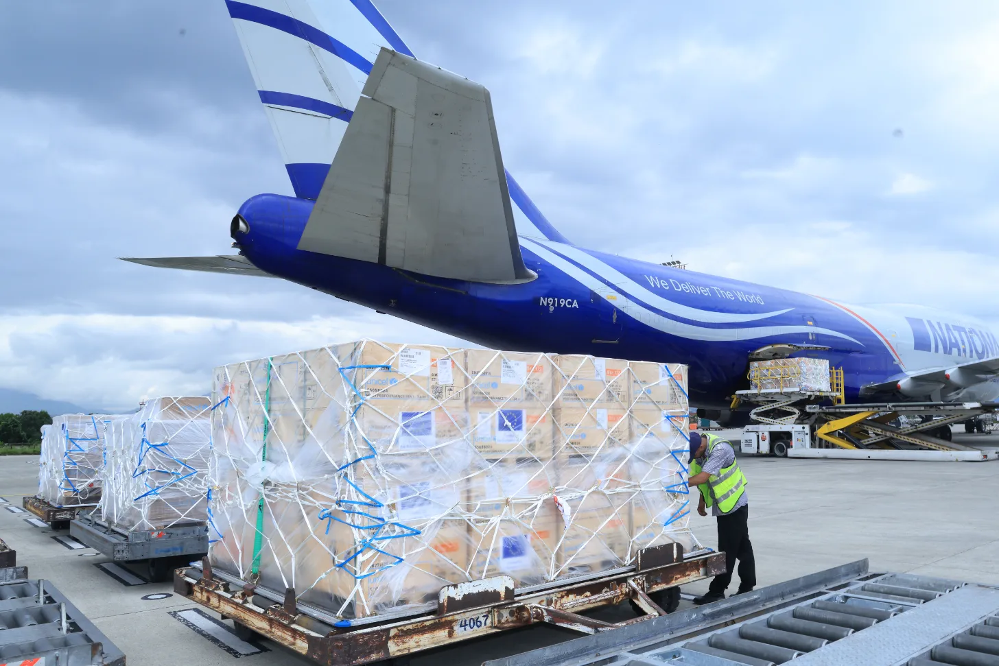 A cargo aircraft with blue and white livery is loaded with wrapped pallets of freight on a tarmac. A ground worker in a high-visibility vest stands near the cargo containers being prepared for loading.