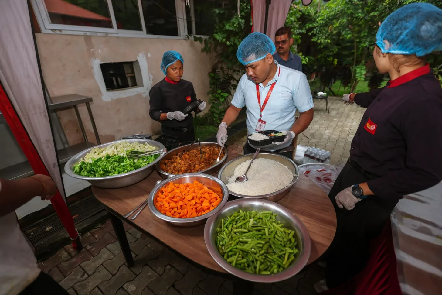 A man wearing a light blue shirt and red ID badge serves food from metal bowls containing green beans, orange carrots, white rice, brown meat dish, and shredded green vegetables to people wearing blue hairnets and dark uniforms in an outdoor courtyard setting.