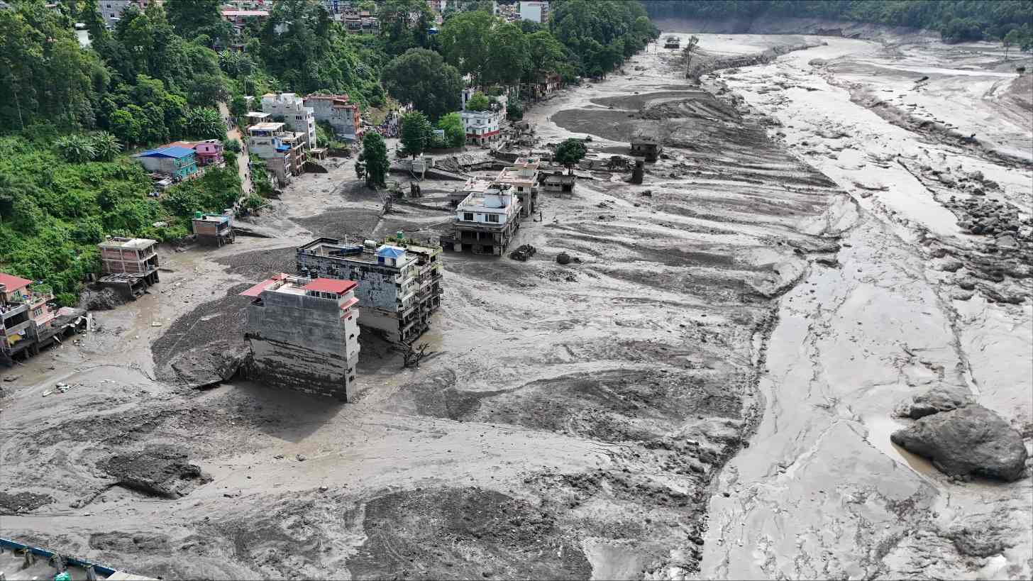 A riverbed filled with gray sediment and water flows past multi-story buildings on a hillside covered with dense green vegetation. The exposed riverbank shows erosion patterns and debris scattered across the pale ground.
