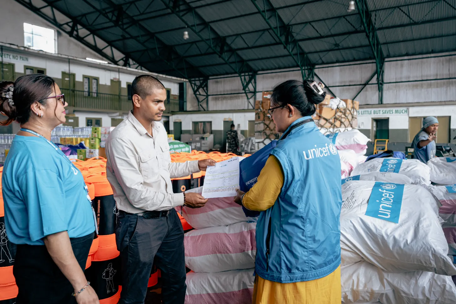 A man in a light gray shirt hands a document to a woman wearing a blue UNICEF vest in a warehouse. Orange and white packaged items are stacked nearby, with other UNICEF workers visible in the background.