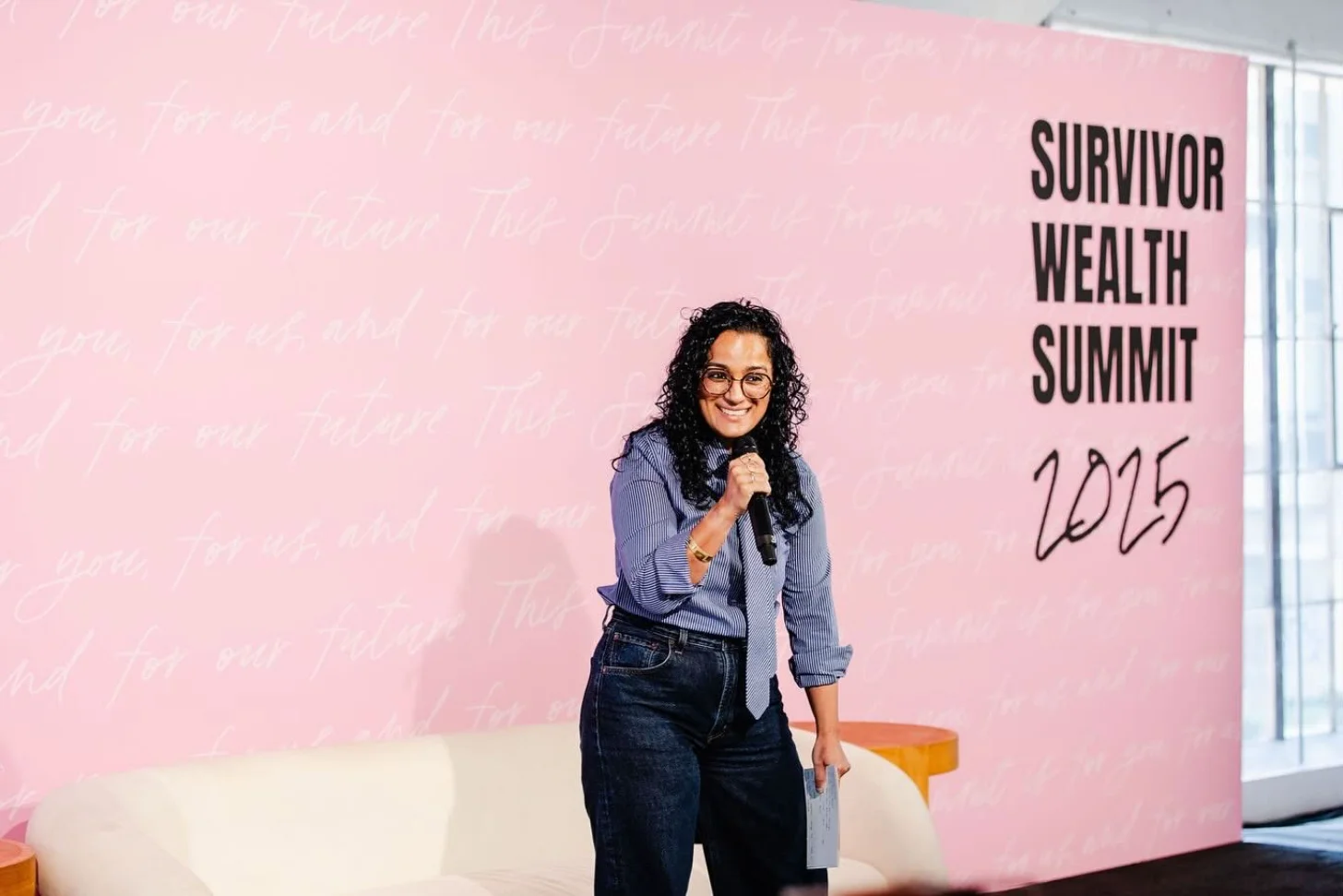 Woman holding a microphone stands in front of a pink wall reading "SURVIVOR WEALTH SUMMIT 2025".