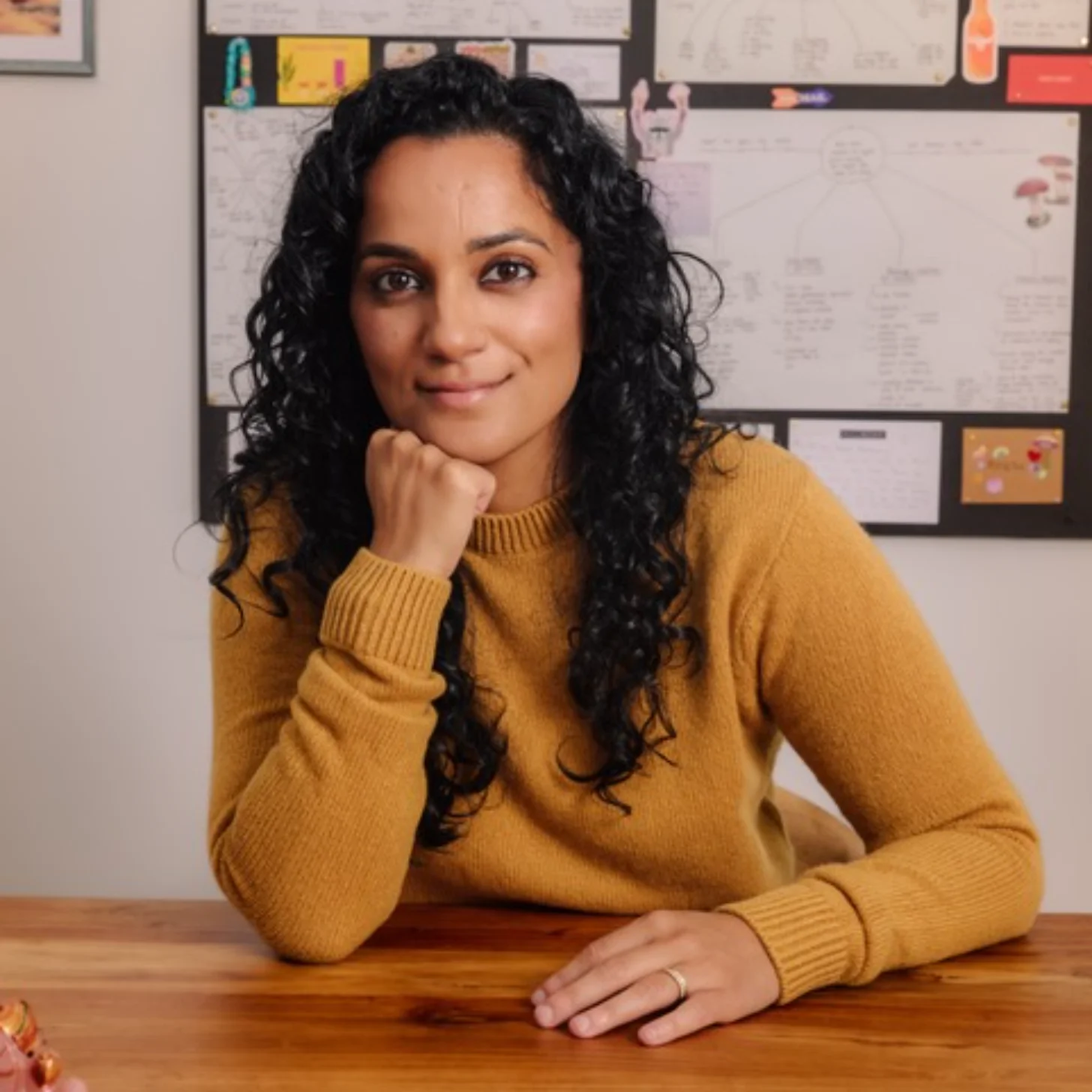 Woman with long black curly hair wearing a mustard yellow ribbed sweater, seated at a wooden table with her chin resting on her hand, facing the camera. A bulletin board with papers and documents is visible behind her.