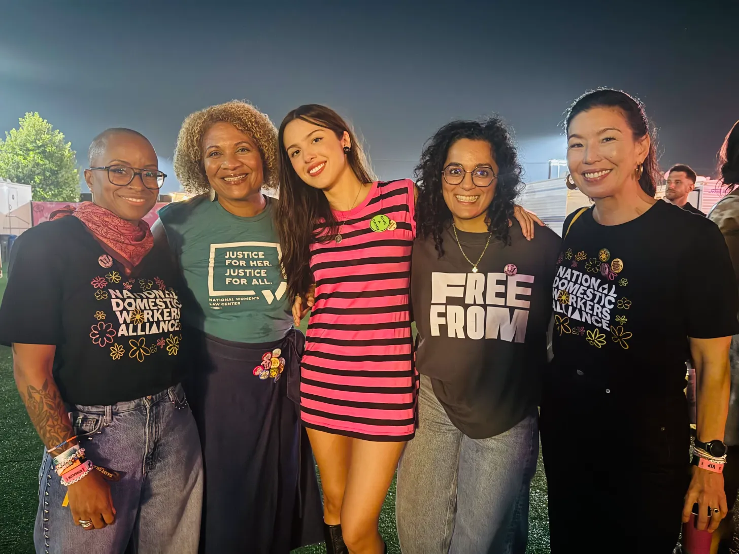 Five people stand together outdoors at dusk, smiling at the camera. They wear t-shirts with text including "National Domestic Workers Alliance," "Justice for Her. Justice for All," and "Free From." In the middle is Olivia Rodrigo.